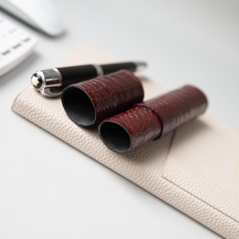 May include: A dark burgundy, crocodile-textured leather pen case consisting of two cylindrical pieces, resting on a beige, pebbled-texture leather mat alongside a black fountain pen with a silver clip and a white star emblem on the cap.