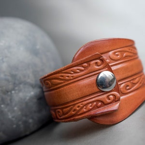 May include: A brown leather bracelet with a decorative pattern and a silver metal snap closure.