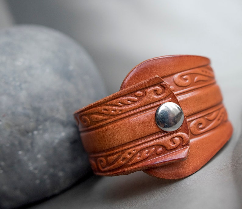 May include: A brown leather bracelet with a decorative pattern and a silver metal snap closure.