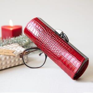 K&ouml;nnte beinhalten: Ein rotes, strukturiertes Brillenetui mit silbernem Verschluss. Das zylindrische Etui liegt neben einer Brille. Eine rote herzf&ouml;rmige Kerze und ein verpacktes Geschenk befinden sich im Hintergrund.