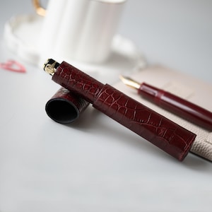 May include: A dark burgundy fountain pen case with a textured crocodile skin pattern. The cylindrical case features a matching removable cap and a gold-toned metal accent at the top. The interior is lined in black, and the case is shown with a fountain pen.