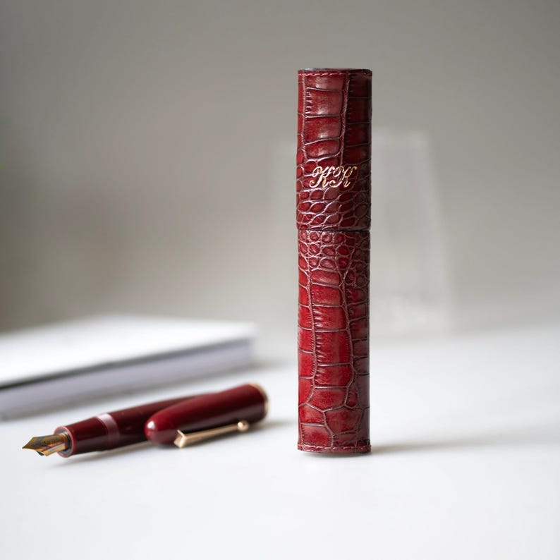 May include: A cylindrical burgundy pen case with a textured crocodile skin pattern and the gold embossed initials KK. Beside the case lies a matching burgundy fountain pen with a gold-colored nib and a gold clip, positioned horizontally.
