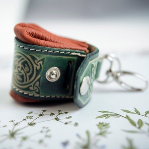 May include: A green leather keychain with a brown leather flap and a Celtic knot design. The keychain has a silver metal ring and a silver metal snap.