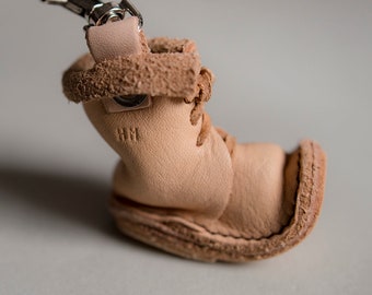 Puede incluir: Un pequeño llavero de cuero marrón claro en forma de bota con un cierre de metal plateado. La bota tiene un diseño de cordones y las letras "HM" están grabadas en el lateral.