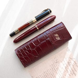 Könnte beinhalten: Zwei Füllfederhalter in einem burgunderfarbenen Lederetui mit Kroko-Muster. Das Etui trägt die Initialen "F.D." in Gold geprägt. Ein Stift ist schwarz mit goldenen Akzenten und der andere ist burgunderfarben.