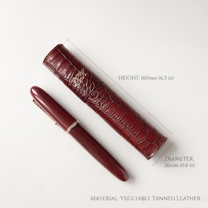 May include: A burgundy fountain pen with a gold-tone band and a matching cylindrical vegetable tanned leather case featuring a crocodile-embossed texture. The case has a height of 6.3 inches and a diameter of 0.8 inches, with gold-tone lettering.