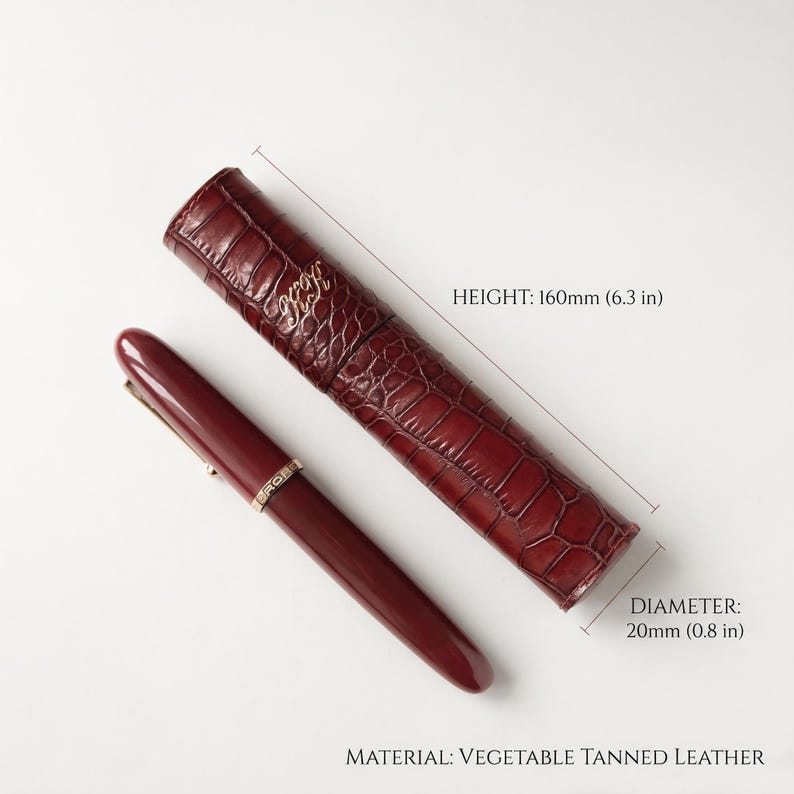 May include: A burgundy fountain pen with a gold-tone band and a matching cylindrical vegetable tanned leather case featuring a crocodile-embossed texture. The case has a height of 6.3 inches and a diameter of 0.8 inches, with gold-tone lettering.