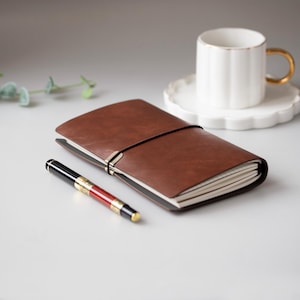 Op de afbeelding: Een bruin leren notitieboek met een zwarte elastische sluiting en een zwart-rode pen. Een witte theekop met een gouden oor en rand staat op een witte schotel. Het notitieboek is open en toont lege pagina's.