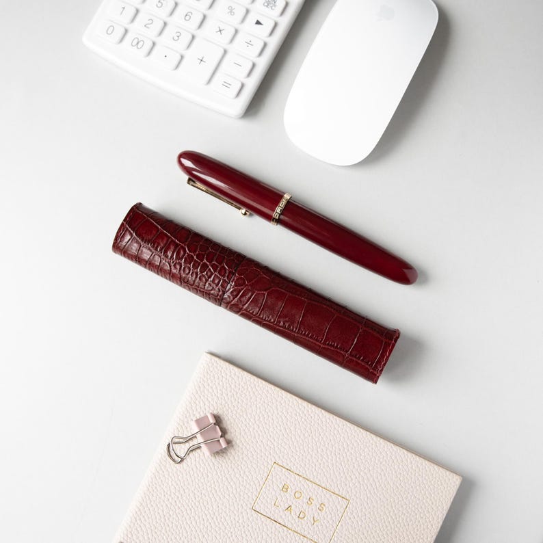 May include: A deep burgundy fountain pen with a gold-toned clip and band, paired with a matching burgundy faux-alligator textured pen case. Below lies a pale pink notebook with the text BOSS LADY embossed in gold and a silver metal binder clip.