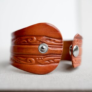 May include: A brown leather cuff bracelet with a decorative embossed pattern and silver-toned snap closures.