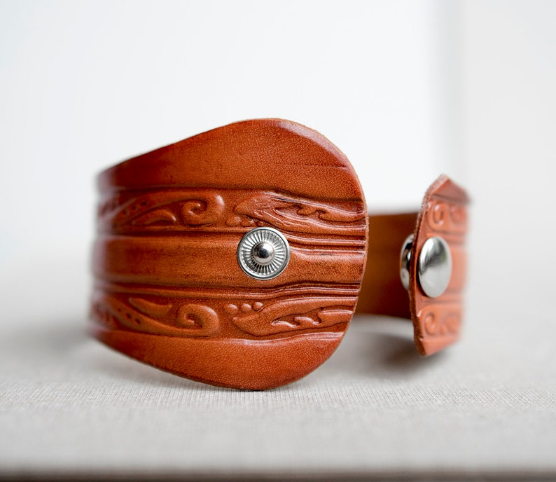 May include: A brown leather cuff bracelet with a decorative embossed pattern and silver-toned snap closures.