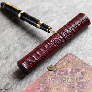 May include: A black fountain pen with gold-colored accents and a metallic nib resting on a cylindrical, deep red, crocodile-textured leather pen case. The case features a glossy finish and a distinct scale pattern across its entire surface.