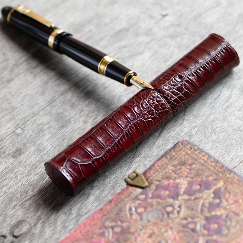May include: A black fountain pen with gold-colored accents and a metallic nib resting on a cylindrical, deep red, crocodile-textured leather pen case. The case features a glossy finish and a distinct scale pattern across its entire surface.