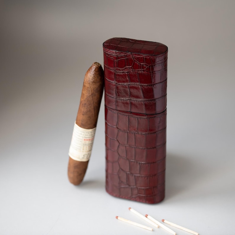 May include: A brown cigar with a white label rests next to a burgundy leather cigar case. The case has a textured, crocodile-like pattern. Three matches are scattered near the case.