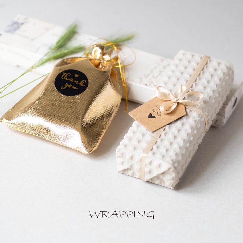 K&ouml;nnte beinhalten: Ein goldener metallischer Geschenkt&uuml;te mit einem schwarzen "Danke"-Aufkleber und einem wei&szlig;en strukturierten Paket, das mit einem Band gebunden ist. Ein kleines braunes Etikett mit einem Herz ist am Paket befestigt. Das Wort "WRAPPING" steht unten.