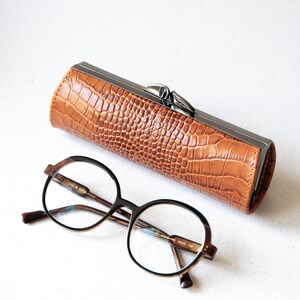 K&ouml;nnte beinhalten: Ein braunes Brillenetui mit Krokodil-Textur und silbernem Verschluss, neben einer Brille mit rundem Rahmen. Die Brille hat einen dunkelbraunen Rahmen mit klaren Gl&auml;sern. Das Etui ist zylindrisch.