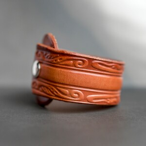 May include: A brown leather bracelet with a decorative embossed design. The bracelet has a silver snap closure.