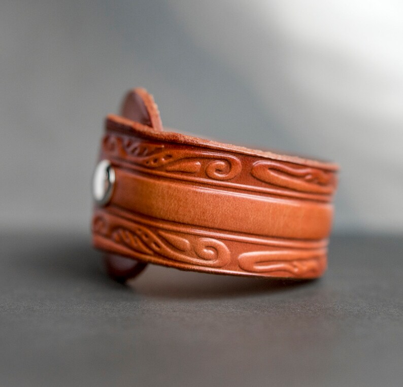 May include: A brown leather bracelet with a decorative embossed design. The bracelet has a silver snap closure.