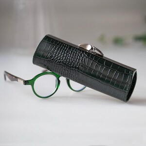 K&ouml;nnte beinhalten: Ein dunkelgr&uuml;nes, strukturiertes Brillenetui mit silbernem Verschluss, neben einer Brille mit gr&uuml;nem Rahmen. Das Etui hat ein Krokodilleder-Muster. Die Brille hat runde Gl&auml;ser und braune und silberne B&uuml;gel.