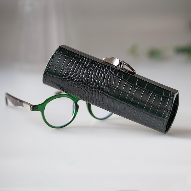 K&ouml;nnte beinhalten: Ein dunkelgr&uuml;nes, strukturiertes Brillenetui mit silbernem Verschluss, neben einer Brille mit gr&uuml;nem Rahmen. Das Etui hat ein Krokodilleder-Muster. Die Brille hat runde Gl&auml;ser und braune und silberne B&uuml;gel.