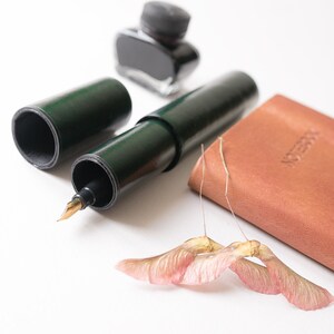 May include: A green leather pen case with a gold nib fountain pen, a small bottle of black ink, and a brown leather notebook with the word "Notebook" embossed on the cover.