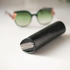 K&ouml;nnte beinhalten: Ein dunkelgr&uuml;nes, zylindrisches Brillenetui mit einem strukturierten, krokodilartigen Muster. Das Etui hat einen silberfarbenen Metallverschluss. Im Hintergrund eine Sonnenbrille mit gr&uuml;nem Farbverlauf und goldfarbenen Akzenten.