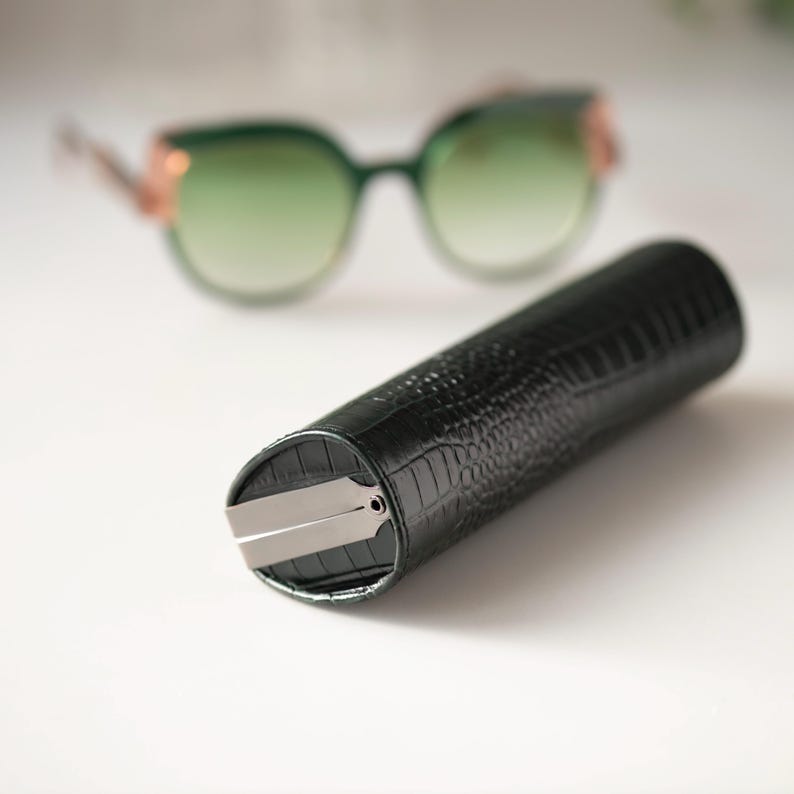 K&ouml;nnte beinhalten: Ein dunkelgr&uuml;nes, zylindrisches Brillenetui mit einem strukturierten, krokodilartigen Muster. Das Etui hat einen silberfarbenen Metallverschluss. Im Hintergrund eine Sonnenbrille mit gr&uuml;nem Farbverlauf und goldfarbenen Akzenten.
