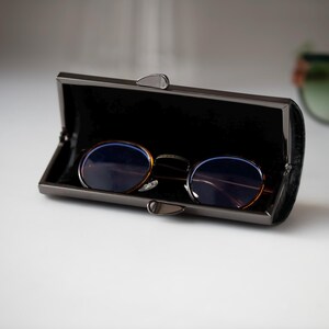 K&ouml;nnte beinhalten: Eine runde Brille mit braunem Gestell und blauen Gl&auml;sern liegt in einem offenen, dunkelgrauen Brillenetui. Das Etui hat einen Schnappverschluss und ein schwarzes Innenfutter. Das Brillenetui befindet sich auf einer wei&szlig;en Oberfl&auml;che.