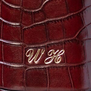 May include: A deep burgundy item featuring a textured crocodile skin pattern. The surface is embossed with the gold-colored cursive initials WH, which are centered and clearly visible against the dark, glossy, and patterned background.