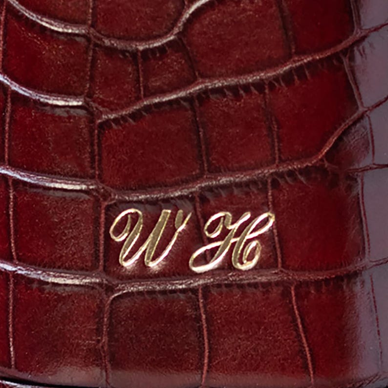 May include: A deep burgundy item featuring a textured crocodile skin pattern. The surface is embossed with the gold-colored cursive initials WH, which are centered and clearly visible against the dark, glossy, and patterned background.