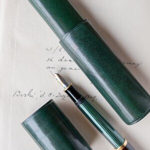 May include: A green leather pen case with a matching green and gold fountain pen. The pen case is closed and the pen is lying on top of it.