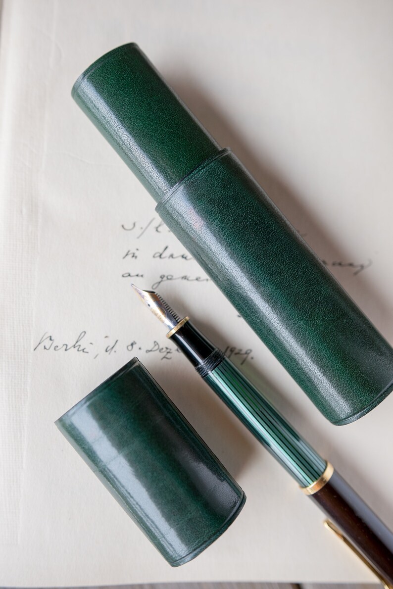 May include: A green leather pen case with a matching green and gold fountain pen. The pen case is closed and the pen is lying on top of it.