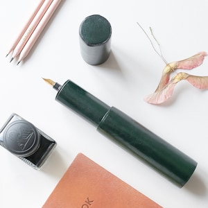 May include: A dark green leather pen case with a gold nib fountain pen. The pen case is lying on a white surface with a brown leather notebook, a bottle of ink, and a few dried maple seeds.
