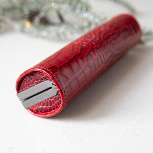 K&ouml;nnte beinhalten: Ein rotes, zylindrisches Etui mit einem strukturierten, krokodilartigen Muster. Das Etui hat einen silberfarbenen Verschluss und N&auml;hte. Das Etui ist f&uuml;r eine Schere oder &auml;hnliche Werkzeuge konzipiert.