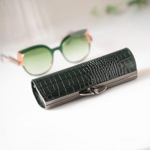 K&ouml;nnte beinhalten: Ein dunkelgr&uuml;nes, rechteckiges Brillenetui mit einer strukturierten, krokodilartigen Pr&auml;gung und einem silberfarbenen Verschluss. Im Hintergrund befindet sich eine Sonnenbrille mit gr&uuml;nem Farbverlauf und goldfarbenen Akzenten. Das Etui dient zum Schutz von Brillen.