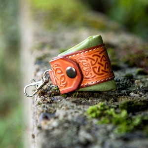 May include: A small, green leather pouch with a brown leather flap and a Celtic knot design. The pouch has a silver key ring attached.