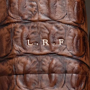 May include: A dark brown leather item with a textured, scale-like pattern resembling alligator skin. The surface features the embossed capital letters L. R. F. in a light, metallic-toned font centered horizontally across the middle of the material.