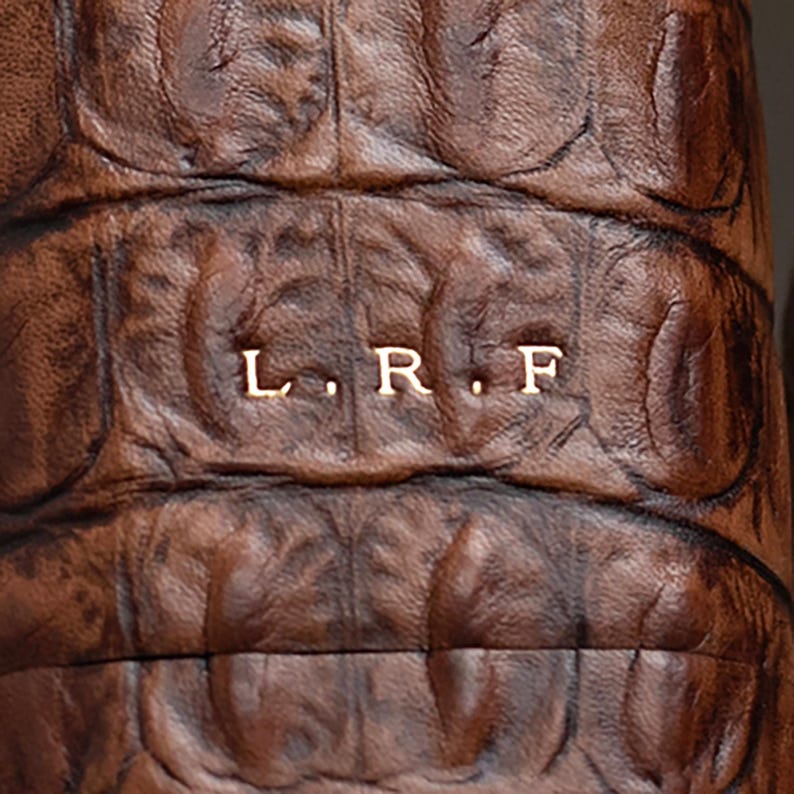 May include: A dark brown leather item with a textured, scale-like pattern resembling alligator skin. The surface features the embossed capital letters L. R. F. in a light, metallic-toned font centered horizontally across the middle of the material.