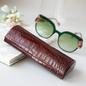 Puede incluir: Un estuche marrón texturizado para gafas con un patrón de cocodrilo. Gafas de sol con tinte verde con montura verde oscuro y detalles dorados cerca. El estuche es rectangular y está cerrado.