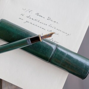 May include: A green leather pen case with a gold-toned fountain pen inside. The pen has a green and gold striped barrel and a gold-toned nib. The case is lying on a white piece of paper with handwritten text that reads "J/L. Herz. Jue in dankbarer Erinnerung an unseren ersten"