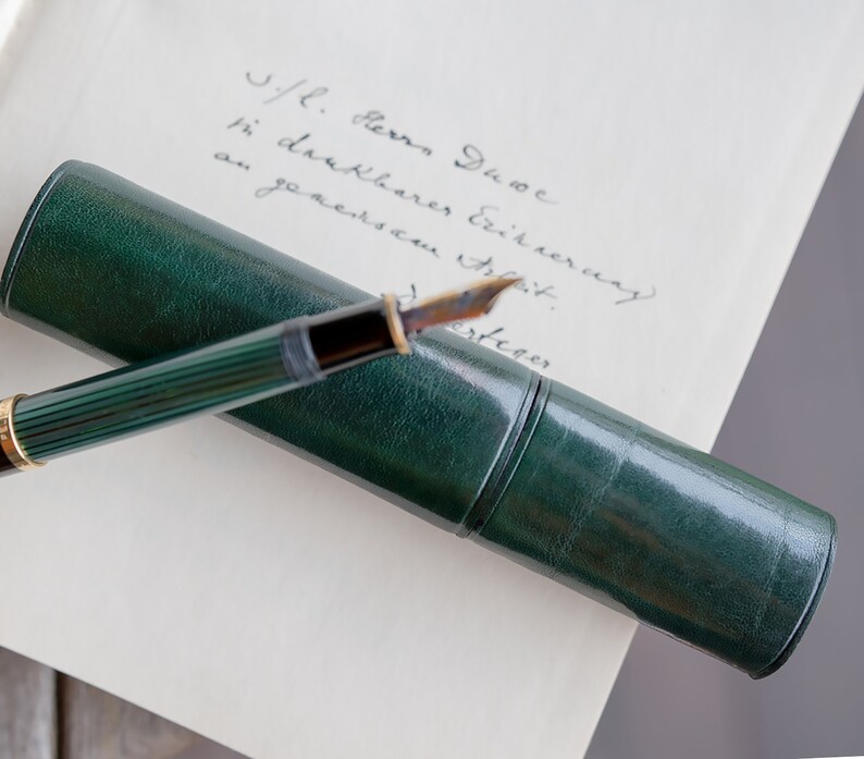 May include: A green leather pen case with a gold-toned fountain pen inside. The pen has a green and gold striped barrel and a gold-toned nib. The case is lying on a white piece of paper with handwritten text that reads "J/L. Herz. Jue in dankbarer Erinnerung an unseren ersten"