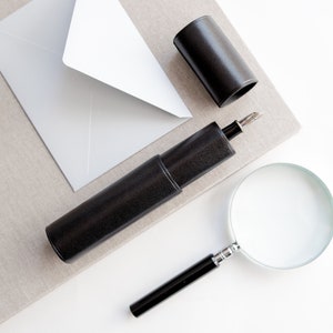 Può includere: Un astuccio per penne in pelle nera con una penna argentata, un tubo in pelle nera e una lente d'ingrandimento su una superficie beige.