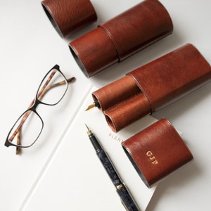 K&ouml;nnte beinhalten: Vier braune Leder-Stift-Etuis, eines mit einem Doppel-Stift-Fach und eines mit den Initialen "CJP" gepr&auml;gt auf der Vorderseite. Ein schwarzer und blauer F&uuml;llfederhalter und eine Brille mit Schildpattgestell sind ebenfalls abgebildet.