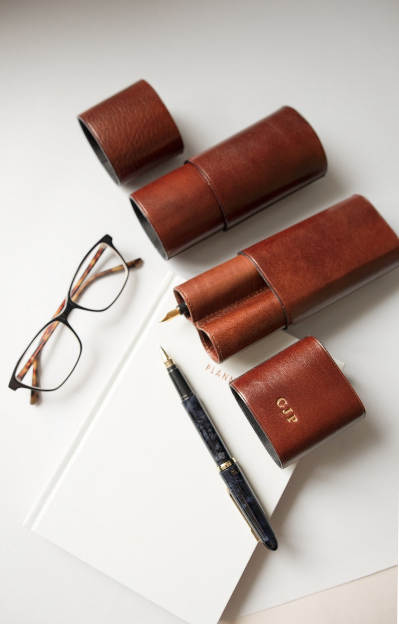 K&ouml;nnte beinhalten: Vier braune Leder-Stift-Etuis, eines mit einem Doppel-Stift-Fach und eines mit den Initialen "CJP" gepr&auml;gt auf der Vorderseite. Ein schwarzer und blauer F&uuml;llfederhalter und eine Brille mit Schildpattgestell sind ebenfalls abgebildet.
