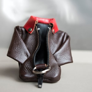 May include: A brown leather coin purse shaped like a jacket with a red collar and a zipper closure.