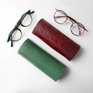 Peut inclure: Vue aérienne de deux paires de lunettes et de deux étuis à lunettes. Une paire a des montures vertes, l'autre des montures écaille. Les étuis sont bordeaux et verts, avec un motif texturé en forme de reptile.
