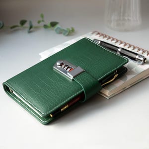 Op de afbeelding: Een groen leren notitieboek met een combinatieslot. Het notitieboek heeft een textuur en witte stiksels. Een pen zit in de penlus. Een spiraalgebonden notitieboek en pen staan op de achtergrond. Het notitieboek is ongeveer 20 cm lang.