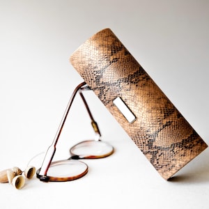 Op de afbeelding: Een bruin en zwart slangenleer-gepatenteerde bril etui met een zilveren sluiting. Het etui ligt op een wit oppervlak met een paar bruine brillen ernaast.