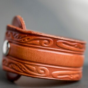May include: A brown leather cuff bracelet with a decorative embossed design. The bracelet has a silver snap closure.