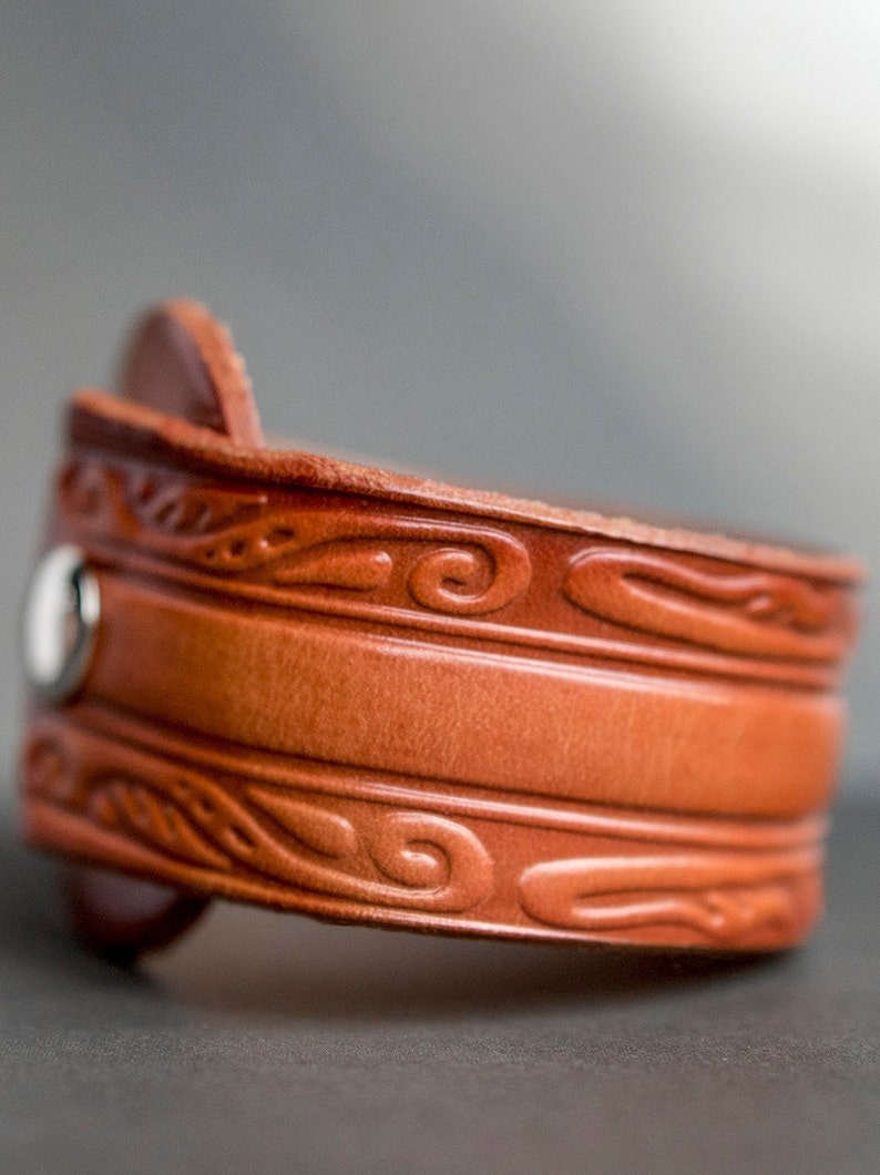 May include: A brown leather cuff bracelet with a decorative embossed design. The bracelet has a silver snap closure.